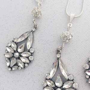 May include: A silver necklace and earrings set with crystal teardrop and round accents. The necklace has a delicate chain and the earrings have a clip-on closure.