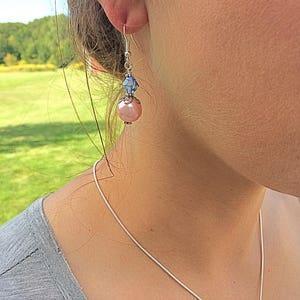 Puede incluir: Un par de pendientes colgantes con una perla rosa y un cristal azul. Los pendientes son de color plateado.