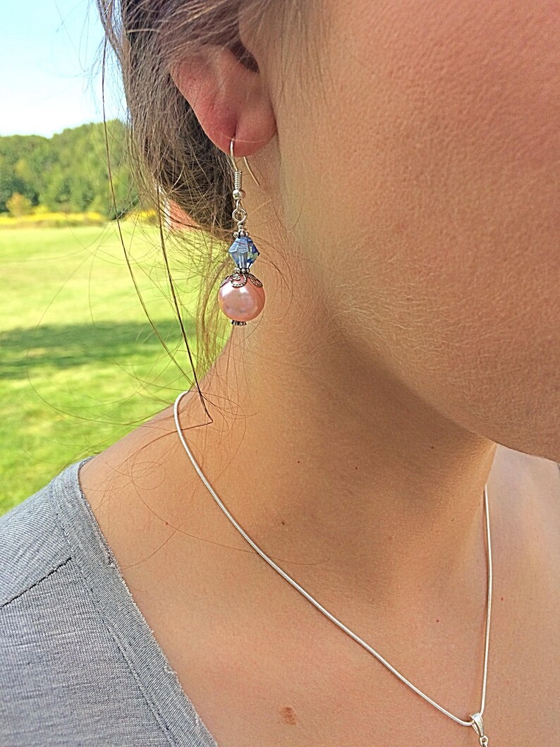 Puede incluir: Un par de pendientes colgantes con una perla rosa y un cristal azul. Los pendientes son de color plateado.