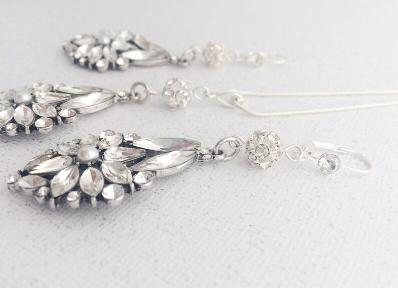 May include: A silver necklace and earrings set with crystal and pearl accents. The necklace features a round crystal pendant and the earrings have a teardrop shape.