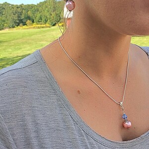 Puede incluir: Un collar de cadena de plata con un colgante de perla rosa y cristal azul. El collar se combina con pendientes con una perla rosa y un cristal azul.