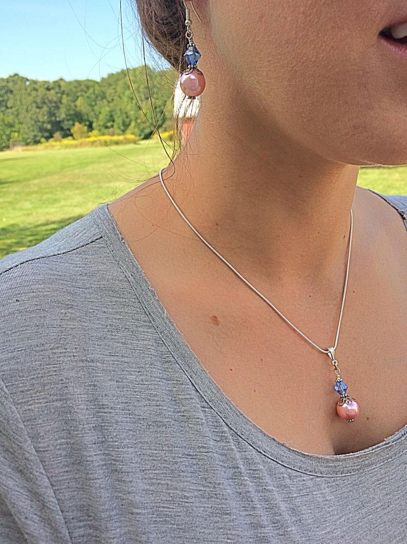 Puede incluir: Un collar de cadena de plata con un colgante de perla rosa y cristal azul. El collar se combina con pendientes con una perla rosa y un cristal azul.