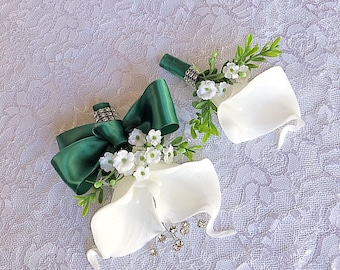 Puede incluir: Dos boutonnières de lirio blanco con cinta verde y flores blancas. Las boutonnières están sobre un fondo de encaje blanco.