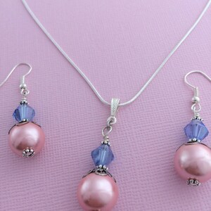 Puede incluir: Un collar de cadena de plata con un colgante de perla rosa y cristal azul, a juego con pendientes de perlas rosas y cristales azules.