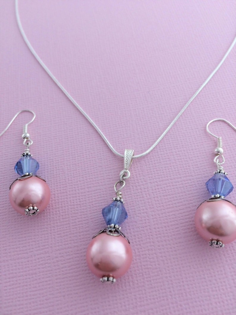 Puede incluir: Un collar de cadena de plata con un colgante de perla rosa y cristal azul, a juego con pendientes de perlas rosas y cristales azules.