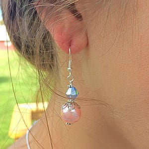 Puede incluir: Un par de pendientes colgantes con una perla rosa y un cristal azul. Los pendientes son de plata y tienen un dise&ntilde;o delicado.