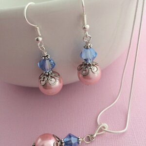 Puede incluir: Un collar de cadena de plata con un colgante de perla rosa y cristal azul. Pendientes a juego con perlas rosas y cristales azules que cuelgan de ganchos de plata.