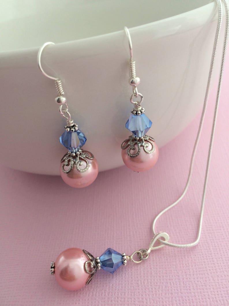 Puede incluir: Un collar de cadena de plata con un colgante de perla rosa y cristal azul. Pendientes a juego con perlas rosas y cristales azules que cuelgan de ganchos de plata.