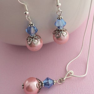 Puede incluir: Un delicado conjunto de collar y pendientes con cuentas de perlas rosas, detalles de cristal azul y filigrana plateada.