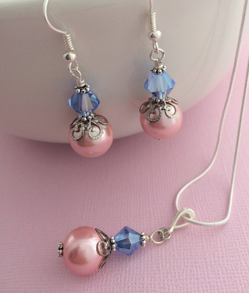 Puede incluir: Un delicado conjunto de collar y pendientes con cuentas de perlas rosas, detalles de cristal azul y filigrana plateada.