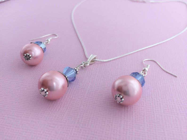 Puede incluir: Un delicado collar de plata y pendientes a juego con una perla rosa y un acento de cristal azul. El conjunto es perfecto para a&ntilde;adir un toque de elegancia a cualquier atuendo.