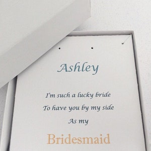 Puede incluir: Una caja blanca con una tarjeta blanca en su interior. La tarjeta tiene el nombre "Ashley" escrito en cursiva azul. La tarjeta tambi&eacute;n tiene el texto "I'm such a lucky bride to have you by my side as my Bridesmaid" escrito en azul y naranja.