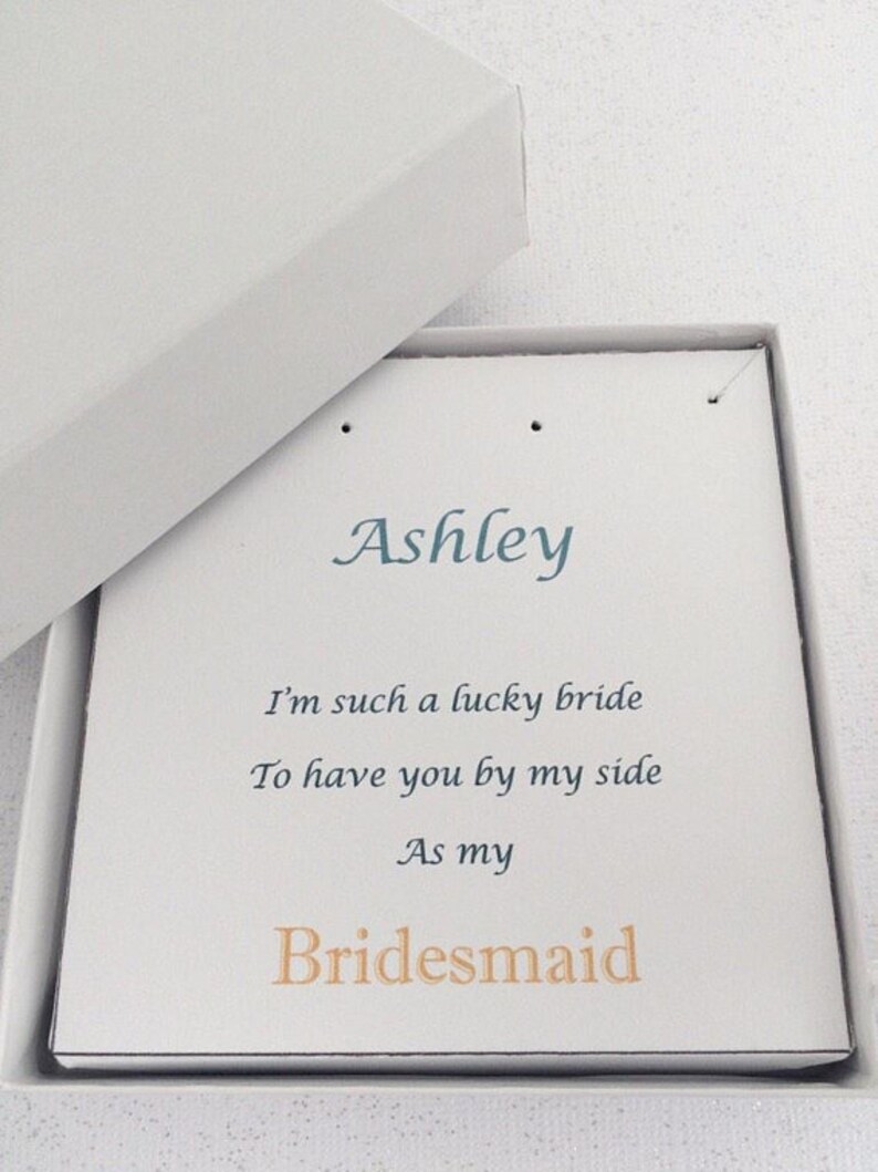 Puede incluir: Una caja blanca con una tarjeta blanca en su interior. La tarjeta tiene el nombre "Ashley" escrito en cursiva azul. La tarjeta tambi&eacute;n tiene el texto "I'm such a lucky bride to have you by my side as my Bridesmaid" escrito en azul y naranja.