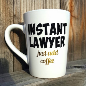 May include: White ceramic coffee mug with black text that reads "INSTANT LAWYER just add coffee". The text "just add coffee" is in gold.