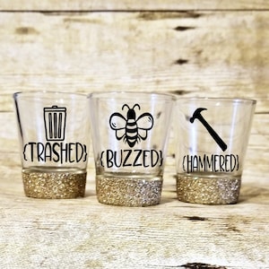 May include: Three clear shot glasses with gold glitter rims. Each glass has a different graphic and text: "Trashed" with a trash can, "Buzzed" with a bee, and "Hammered" with a hammer.