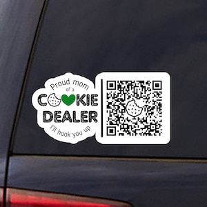 May include: White sticker with black text that reads "Proud mom of a cookie dealer I'll hook you up" with a green heart and a cookie graphic. A black and white QR code is on the right side of the sticker.