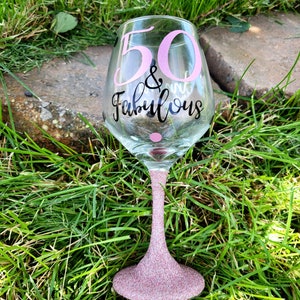 May include: A pink glitter wine glass with the text "50 & Fabulous" in black lettering.