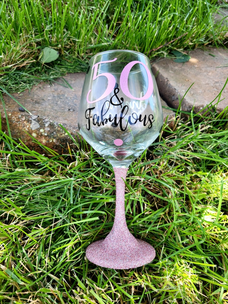 May include: A pink glitter wine glass with the text "50 & Fabulous" in black lettering.