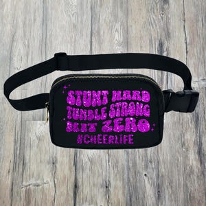 May include: Black fanny pack with a zippered main compartment and adjustable strap. The front features the text "STUNT HARD TUMBLE STRONG HIT ZERO #CHEERLIFE" in sparkly purple lettering. The bag is designed for cheerleaders.