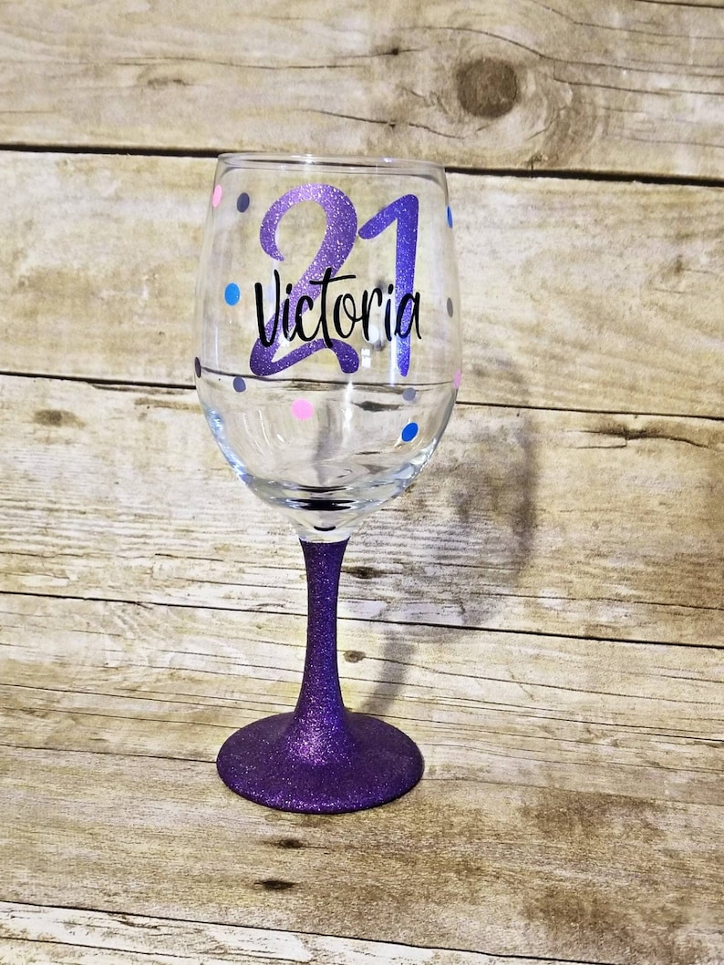May include: Purple glitter wine glass with a white design that says "21 Victoria" with colorful polka dots.