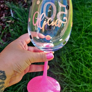 May include: A pink stemless wine glass with black polka dots and gold glitter. The glass has the number "30" and the name "Alicia" written on it.