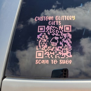 Pode incluir: Um adesivo rosa e branco na janela de um carro que diz "Custom Glittery Gifts" e tem um c&oacute;digo QR com um &iacute;cone de cupcake. O texto abaixo do c&oacute;digo QR diz "Scan to Shop".