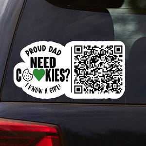 May include: A white car decal with the text "PROUD DAD NEED COOKIES? I KNOW A GIRL!" and a QR code. The words are in black, with a green heart and a cookie graphic. The decal is on a car window.