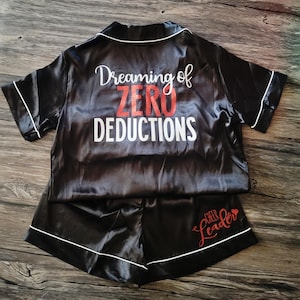May include: Black satin pajama set with short sleeves and shorts. The top has the text "Dreaming of ZERO DEDUCTIONS" in white and red glitter. The shorts have the text "CHEER LEADER" in red glitter.