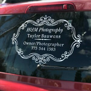 May include: White vinyl decal with a decorative frame and the text "H&M Photography, Taylor Bauwens, Owner/Photographer, 775 544 1383" on a red car window.