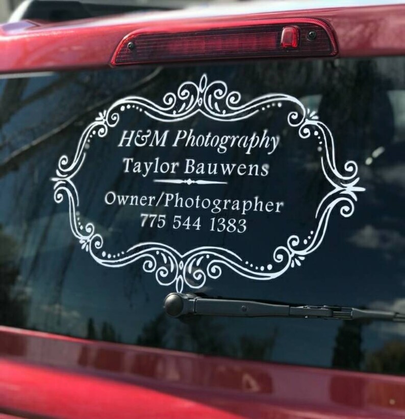 May include: White vinyl decal with a decorative frame and the text "H&M Photography, Taylor Bauwens, Owner/Photographer, 775 544 1383" on a red car window.