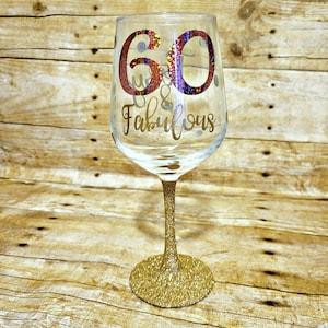 May include: A gold glitter wine glass with the text "60 & Fabulous" in gold glitter lettering. The number 60 is in red glitter with iridescent accents.