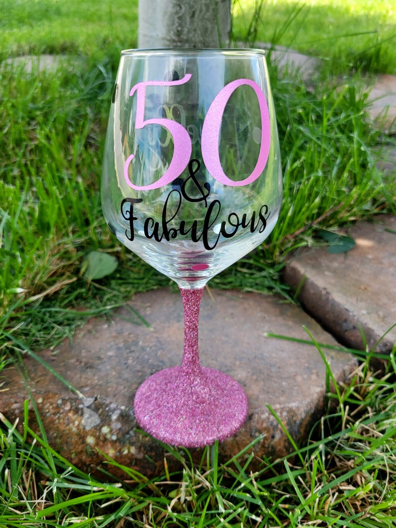 May include: A pink glitter wine glass with the text "50 & Fabulous" in black lettering. The number 50 is in pink.