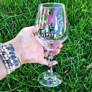 May include: A clear glass wine glass with black and pink polka dots. The glass has a pink and white glitter design that says "50 & Fabulous".