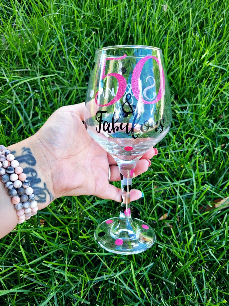 May include: A clear glass wine glass with black and pink polka dots. The glass has a pink and white glitter design that says "50 & Fabulous".