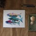 A watercolour print of three Salmon shapes in silhouette form with images of trees, waves and triangles inside. Colours include light blue, teal, pink, orange green and yellow. The background is white. The horizontal print rests on a dark table.