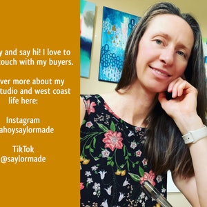 A photo of the artist in her studio. She looks  at the camera and smiles. Behind her is an array of brightly coloured paintings.