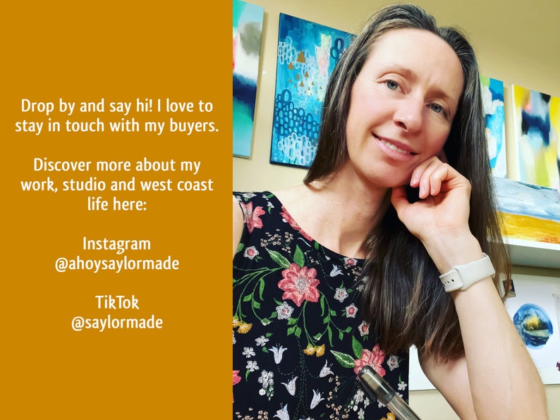 A photo of the artist in her studio. She looks  at the camera and smiles. Behind her is an array of brightly coloured paintings.