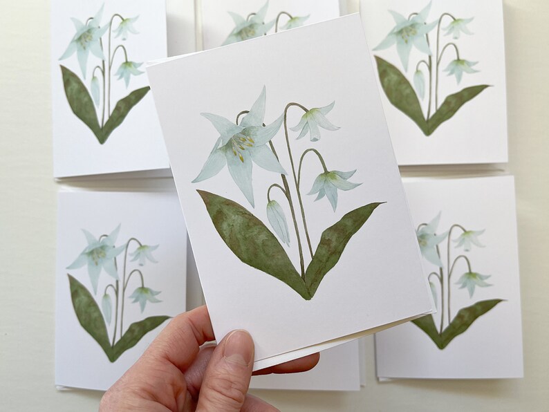 Easter Lily botanical watercolor painting printed onto a white card. Image is centred on a white cardstock background. Close detail of the card in foreground, multiples of same card in background.