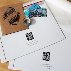 An example of packaging material. Durable cardboard stay-flat mailers have the artists logo stamp displayed as well as social media and website contact information. A card, tape and packing material lays flat on a table display.