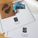 An example of packaging material. Durable cardboard stay-flat mailers have the artists logo stamp displayed as well as social media and website contact information. A card, tape and packing material lays flat on a table display.