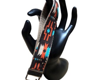 May include: A patterned wrist lanyard with a silver-colored key ring. The lanyard features a geometric design in shades of brown, orange, white, and turquoise. The lanyard is held by a black hand-shaped display.