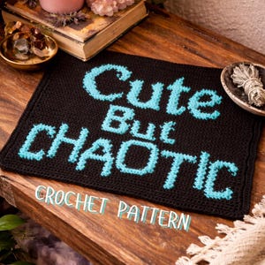 May include: A black crochet square with the words "Cute But Chaotic" in turquoise. The words "CROCHET PATTERN" are below the main text. The square is on a wooden surface with other decorative items.