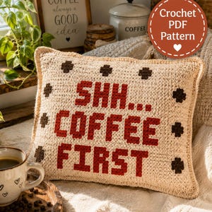 May include: A square crochet cushion in a light beige colour featuring the dark red text SHH... COFFEE FIRST and small dark brown cross-shaped accents. The cushion has a textured yarn surface and a simple border, presented as a crochet PDF pattern.