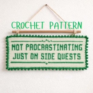 May include: A crocheted sign with the text "NOT PROCRASTINATING JUST ON SIDE QUESTS" in green on an off-white background. The sign is framed with a green border and is suspended by a wooden dowel and twine. The words "CROCHET PATTERN" are above the sign.