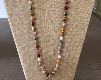 May include: A beaded necklace featuring a variety of round, multi-colored stone beads in shades of brown, tan, cream, and grey, interspersed with smaller metallic spacer beads. A single, textured, metallic leaf-shaped pendant hangs at the center.