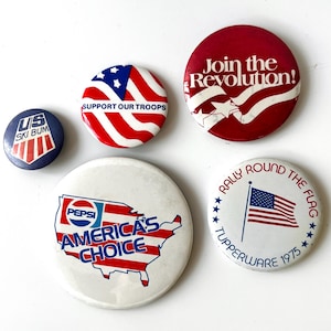 May include: A collection of vintage pinback buttons with patriotic themes. Designs include the American flag, the text "Support Our Troops," and a Pepsi advertisement with the text "America's Choice."