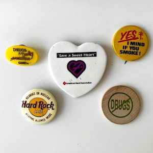 May include: A collection of vintage pins and buttons with various designs and messages. Includes a yellow oval pin with "DRUGS: A Deadly DANGEROUS GAME!" text, a heart-shaped pin with "Save a Sweet Heart", and a Hard Rock Cafe button.