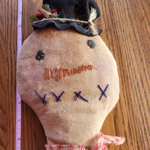 May include: A handmade rag doll with a brown fabric body, a black felt hat with red berries and a brown twine bow, and blue embroidered stitches for eyes and a mouth.