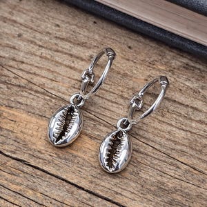 May include: A pair of silver hoop earrings with seashell charms. The earrings are made of shiny metal and feature a detailed seashell design. The earrings are displayed on a wooden surface.