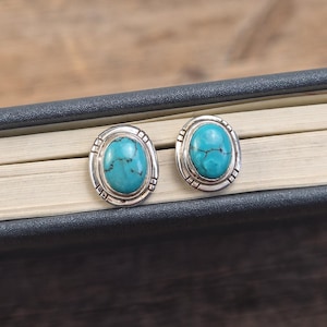 May include: A pair of oval turquoise stud earrings with a silver border. The earrings are set on a dark surface. The turquoise stones have a natural, veined appearance. The earrings are a classic jewellery item.